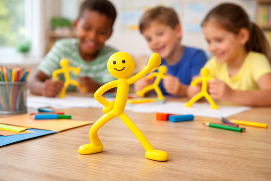 Yellow Stick Man bendable toy standing on a table with children playing in the background, promoting creative and sensory play.